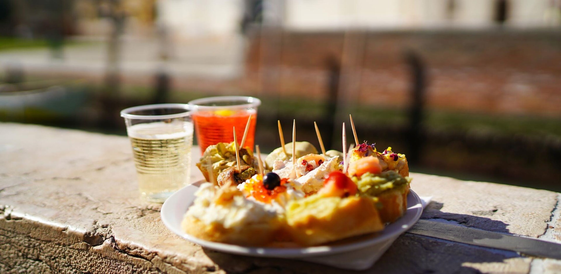 A plate with cicchetti, traditional Venetian snacks and glass of aperol sprits on the background, view on Grand Canal. Travel concept, foodie, food lover. Food blog.