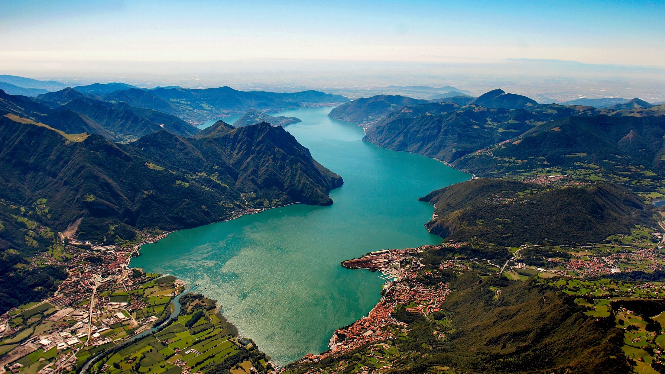 An aerial view of Lake Iseo nestled among the mountains in Italy. The image showcases the lake's emerald green waters surrounded by steep, verdant peaks and picturesque towns along the shoreline.